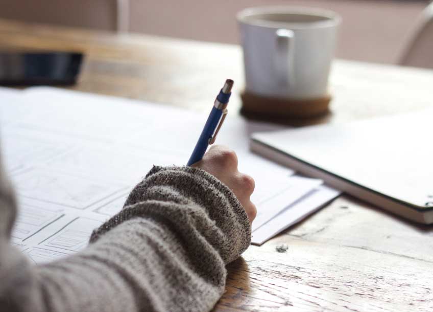 Someone writing on a piece of paper with a cup of coffee on the desk